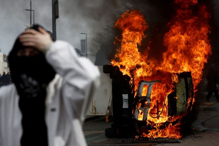 Ένας μασκοφόρος διαδηλωτής στέκεται μπροστά από ένα φλεγόμενο αυτοκίνητο κατά τη διάρκεια συγκρούσεων σε μια διαδήλωση κατά της συνταξιοδοτικής μεταρρύθμισης της γαλλικής κυβέρνησης στη Νάντη, Γαλλία, 28 Μαρτίου 2023.