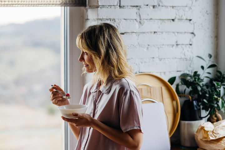 A pensive smiling Caucasian female contemplating while standing near the window and enjoying her healthy meal.