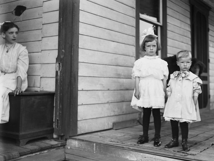 A big sister sits aside as her brother and sister stand for a picture, circa 1903.