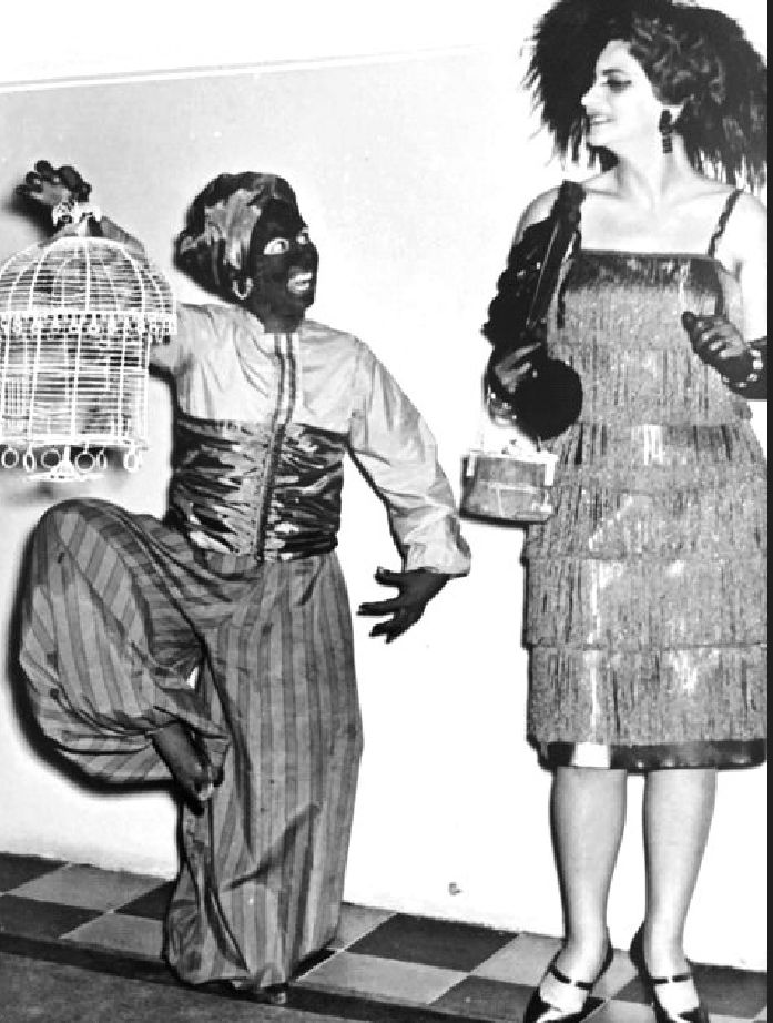 Η συγγραφέας Αθηνά Κακούρη ως έγχρωμος υπηρέτης προαναγγέλλει την άφιξη της Κλάρας Μπόου (Λίνας Δημητροπούλου) σε έναν από τους αποκριάτικους χορούς της Φιλαρμονικής που άφησαν εποχή. 