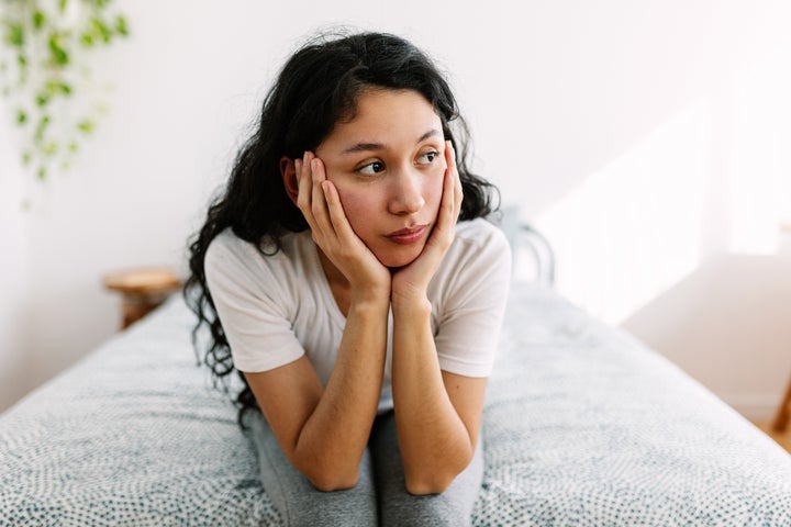 Sad millennial woman sitting on bed looking away thinking about problems and bad feelings