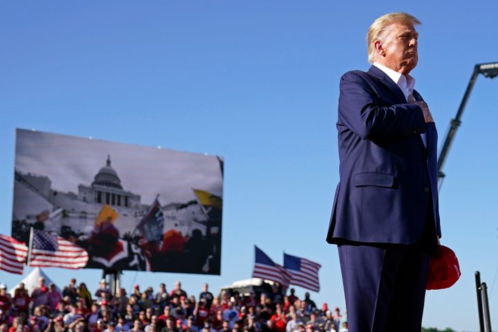 As footage from the Jan. 6, 2021, insurrection at the U.S. Capitol is displayed in the background, former President Donald Trump stands while a song, "Justice for All," is played during a campaign rally on March 25 in Waco, Texas.