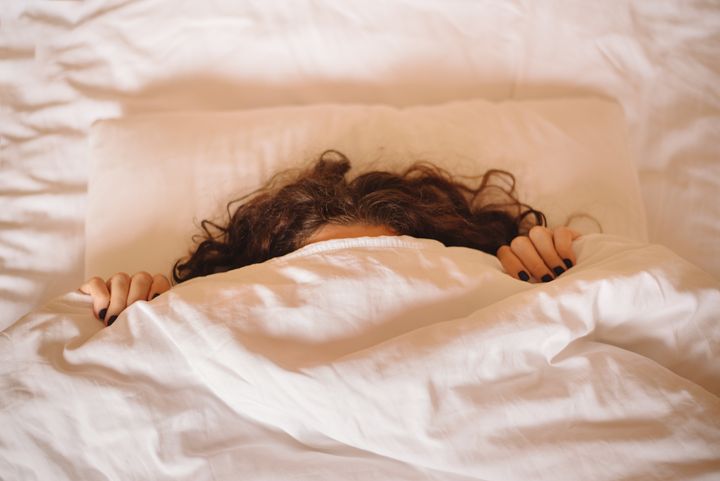 Shy or angry young woman hides under white blanket, only her long brown curly hair and dark manicured hands are visible. Good morning. Girl stretches, raises her hands, and makes plans for day.