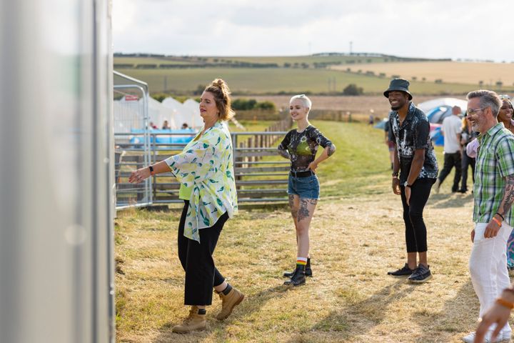A group of festival goers at a festival in Lindisfarne, North East England, standing in a field with the campsite in the background. They are all queuing for the toilets and smiling as one woman is entering a cubicle.