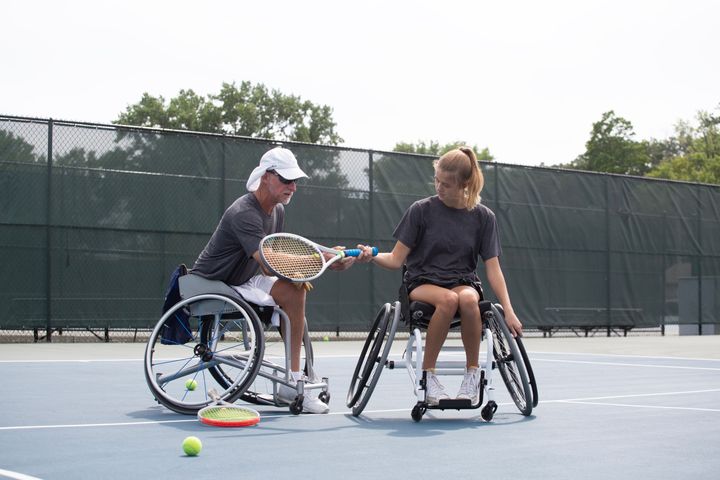 A Caucasian adaptive teenage girl is learning how to play wheel chair tennis with her instructor outside on a summer day, wearing casual athletic clothing.