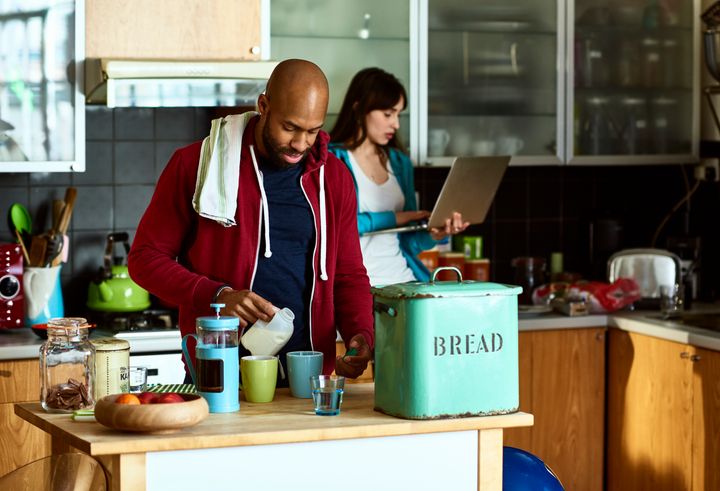 Couple at home on relaxing weekend morning, preparing hot drinks, woman working from home on computer as her partner pours milk into cups