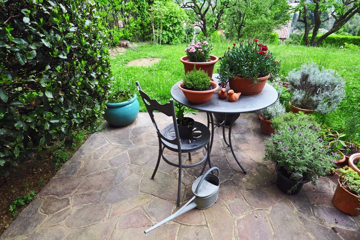 Small paved patio with wrought iron furniture, watering can, various containers with kitchen herbs and flowering plants, lavender, carnation, sedum and other herbal plants against green background. On the left laurel bush.