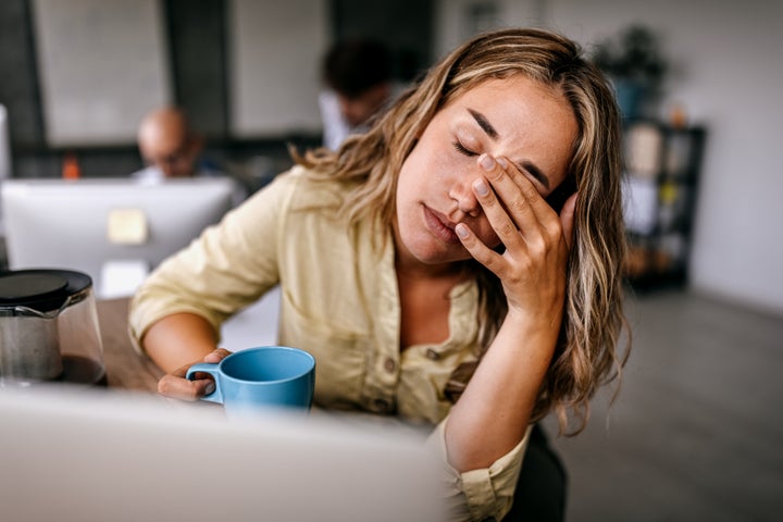 Tired young woman drinking coffee and rubbing her eyes while on a laptop.