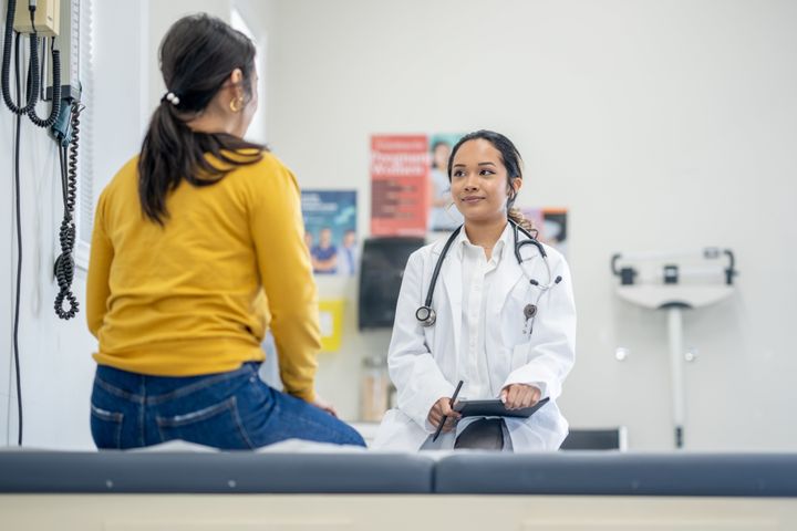 A middle aged woman of Asian decent, sits up on an exam table as her doctor talks with her about her recent test results.