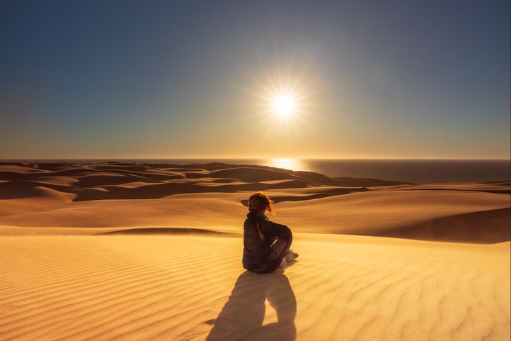 Person sitting on sand in desert with bright sun in the sky