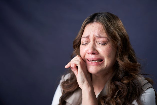 Close up portrait of grieving, crying women in her 50's with eyes closed looking down and hand on chin against blue background