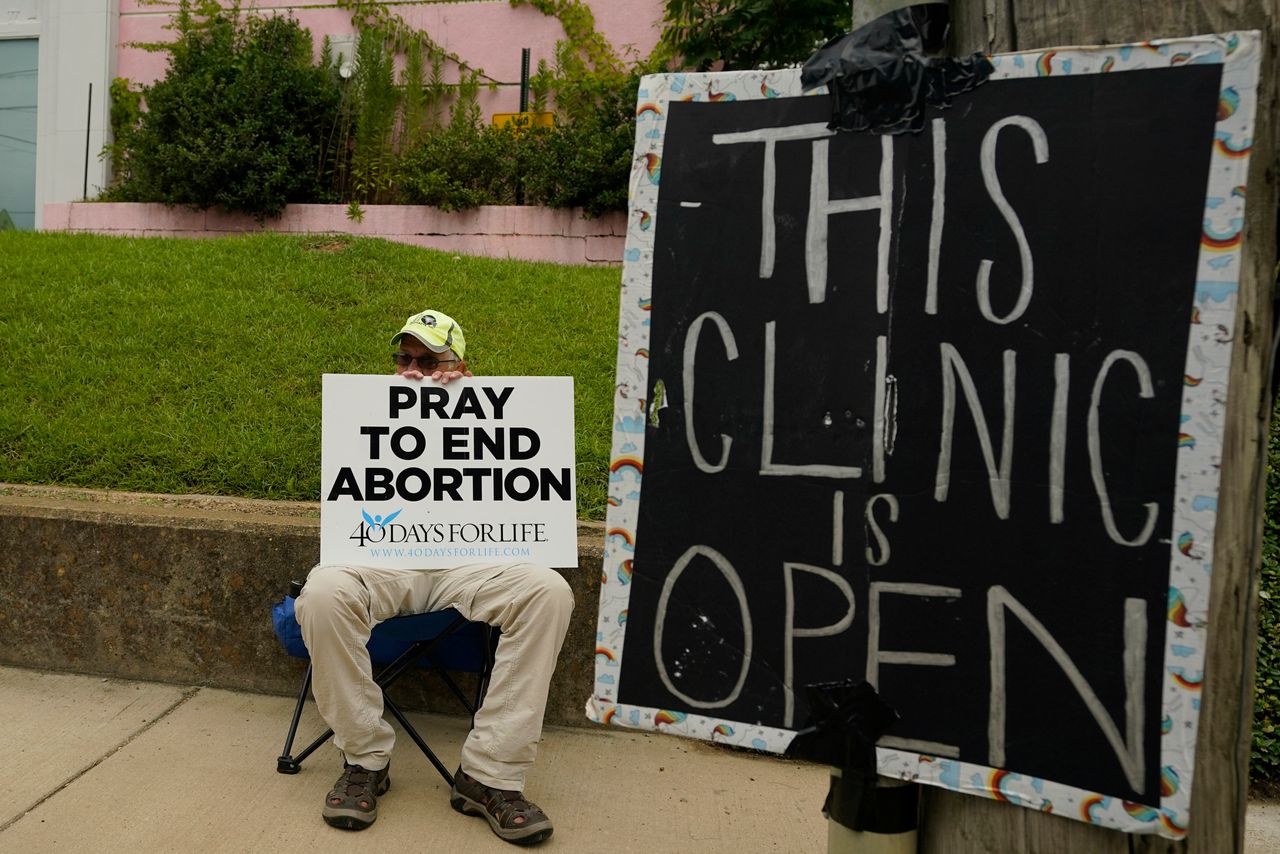 Ahead of the Dobbs decision last year, an anti-abortion supporter sits near a sign stating that Jackson Women's Health Organization clinic is still open in Jackson, Mississippi. Abortion is now completely outlawed in the state.