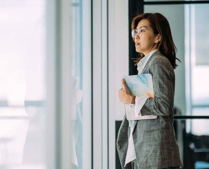 An Asian businesswoman holding digital tablet and look in distance window thinking or planning future success, thoughtful motivated mature businesswoman pondering over new ideas.