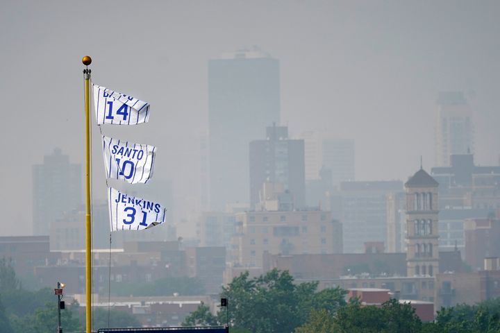 The flags of Chicago Cubs' Hall of Fame players Ernie Banks (14) Ron Santo (10) and Ferguson Jenkins fly off the Wrigley Field left field foul pole as a veil of haze from Canadian wildfires shroud high rise buildings along Lake Michigan before a baseball game between the Cubs and the Philadelphia Phillies Tuesday, June 27, 2023, in Chicago. (AP Photo/Charles Rex Arbogast)
