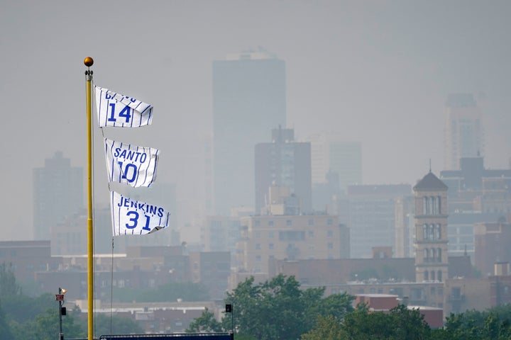 The flags of Chicago Cubs' Hall of Fame players Ernie Banks (14) Ron Santo (10) and Ferguson Jenkins fly off the Wrigley Field left field foul pole as a veil of haze from Canadian wildfires shroud high rise buildings along Lake Michigan before a baseball game between the Cubs and the Philadelphia Phillies Tuesday, June 27, 2023, in Chicago. (AP Photo/Charles Rex Arbogast)