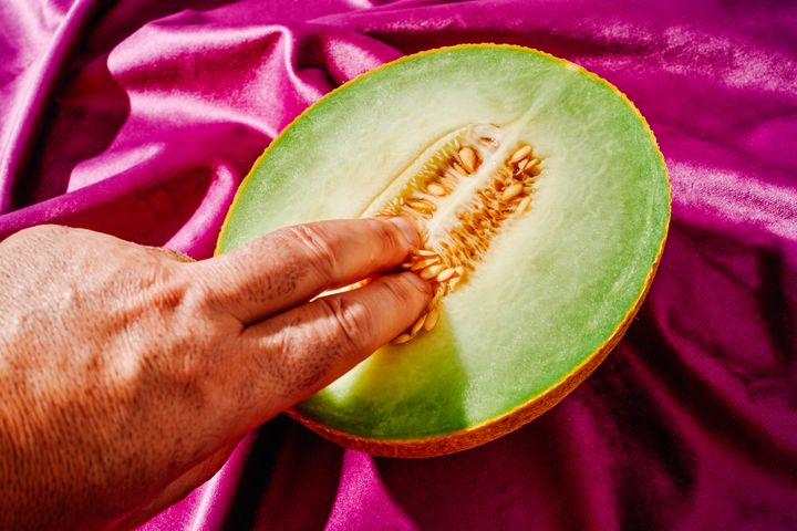 a man is inserting two fingers into the central part of a melon, where the seeds are, placed on a draped purple velvet fabric