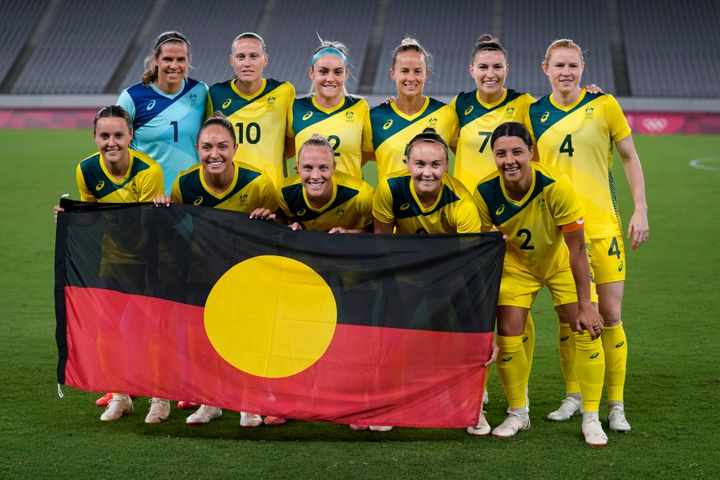 Para pemain sepak bola Australia berfoto bersama dengan bendera Aborigin sebelum pertandingan melawan Selandia Baru di Olimpiade 2021.  Warna bendera melambangkan orang Aborigin dan hubungan mereka dengan tanah, dengan lingkaran kuning melambangkan matahari.