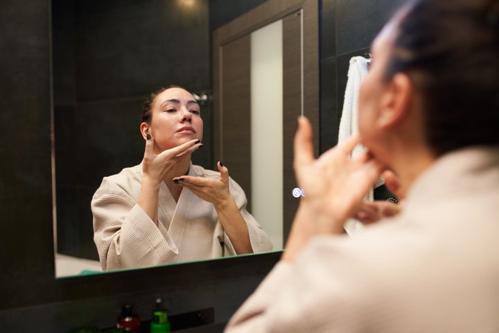 Woman in cotton bathrobe applying cream and massaging chin while looking at mirror during beauty routine in bathroom at home.