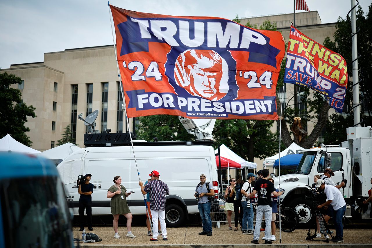 Trump supporters carry a large flag in front of television satellite trucks.