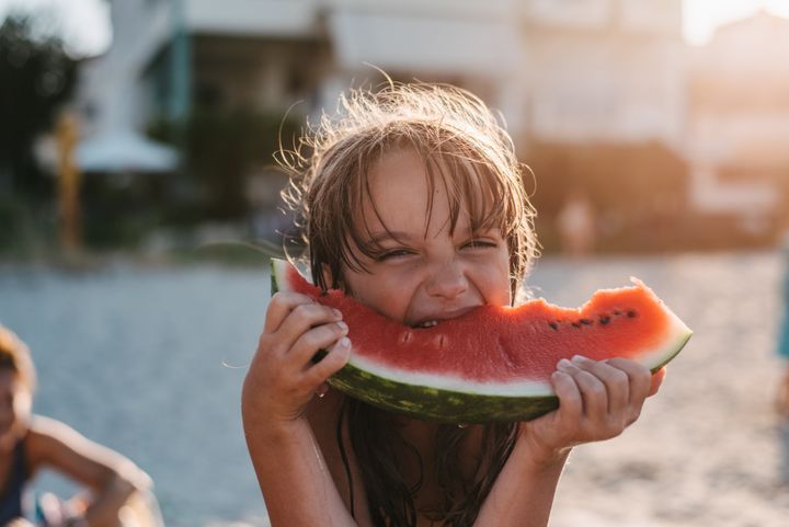 A close-up of a girl on the beach biting on a watermelon looking at the camera squinting