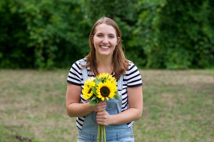 Smiling young woman with brunette hair is wearing jean fabric overalls and a striped shirt while smiling and holding a beautiful bouquet of yellow Sunflowers from the farmers market.