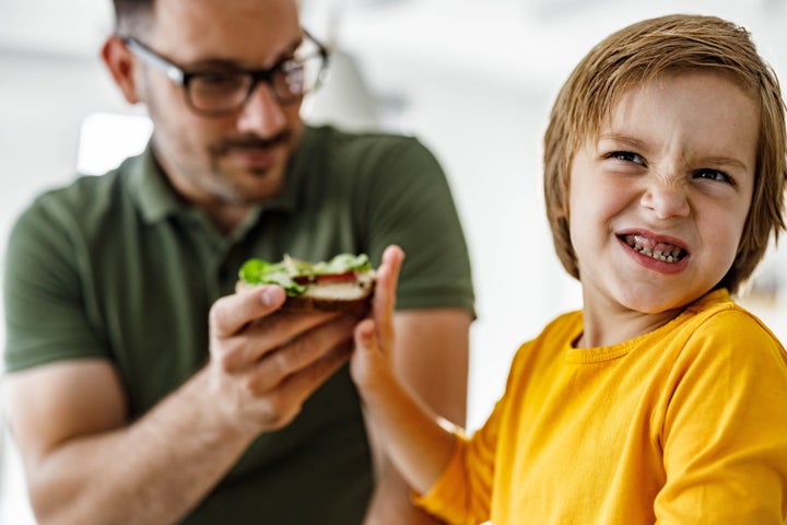 Small boy making a face while refusing to eat a sandwich his dad is giving him.