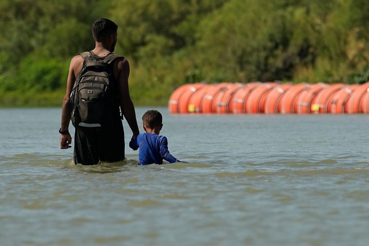 Migrants walk past large buoys being used as a floating border barrier on the Rio Grande on Aug. 1 in Eagle Pass, Texas.