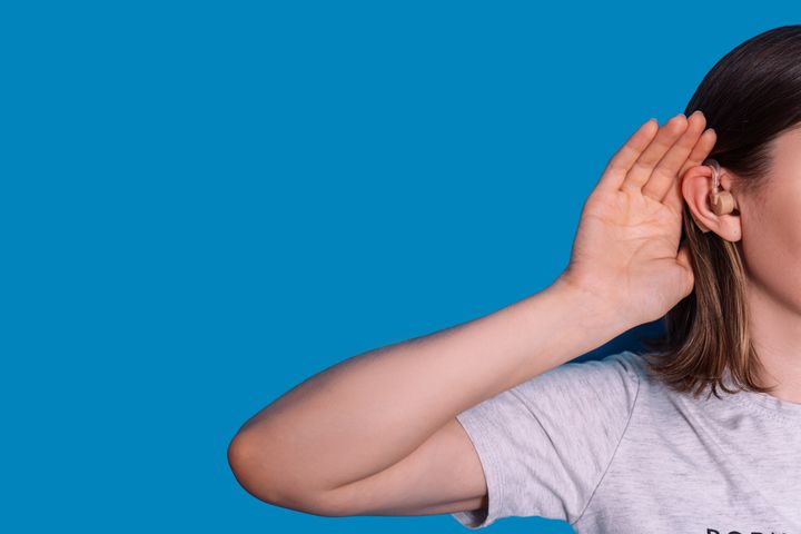 Lady with hearing aid put her hand to her ear against blue background.