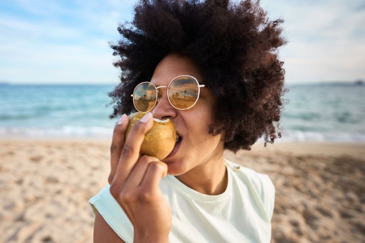Close up portrait of a woman with sunglasses eating a pear on the beach.