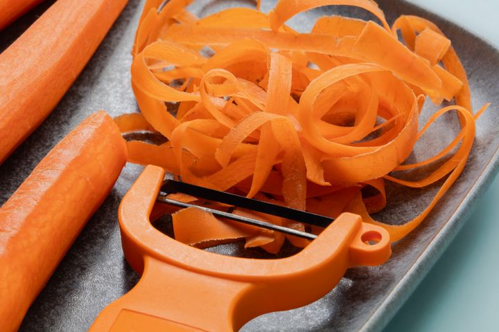 Close view of a blue plate with carrots, carrot peels and a peeler tool on light blue background
