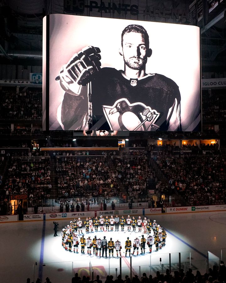 Οι Pittsburgh Penguins και οι Anaheim Ducks συγκεντρώνονται στο κέντρο του πάγου, πριν από έναν αγώνα χόκεϊ NHL στο Πίτσμπουργκ, τη Δευτέρα 30 Οκτωβρίου 2023, για να τιμήσουν τον πρώην παίκτη των Penguin, Adam Johnson, που φαίνεται στον πίνακα, ο οποίος έχασε τη ζωή του σε ένα "φρικτό ατύχημα" ενώ έπαιζε σε αγώνα του αγγλικού πρωταθλήματος χόκεϊ. (AP Photo/Gene J. Puskar)