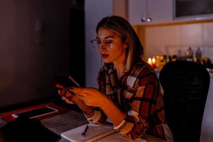 In the time of a blackout caused by an energetic crisis, a woman is working on a laptop while talking over a mobile phone at home under lit candles.