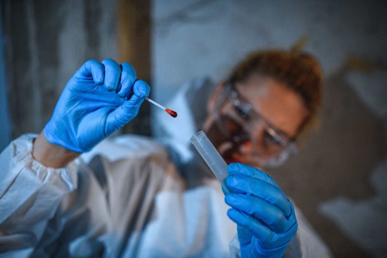 Caucasian woman in a white forensic uniform taking blood samples in a crime scene. She is using professional equipment.