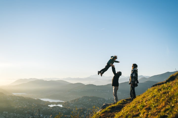 Father throws son in air above head on mountain top at sunset, mother watches