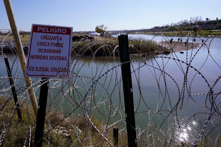 Μετανάστες διασχίζουν το Ρίο Γκράντε προς τις ΗΠΑ από το Μεξικό πίσω από συρματόπλεγμα και μια πινακίδα που προειδοποιεί ότι είναι επικίνδυνη και παράνομη η διέλευση, Τετάρτη 3 Ιανουαρίου 2024, στο Eagle Pass του Τέξας.