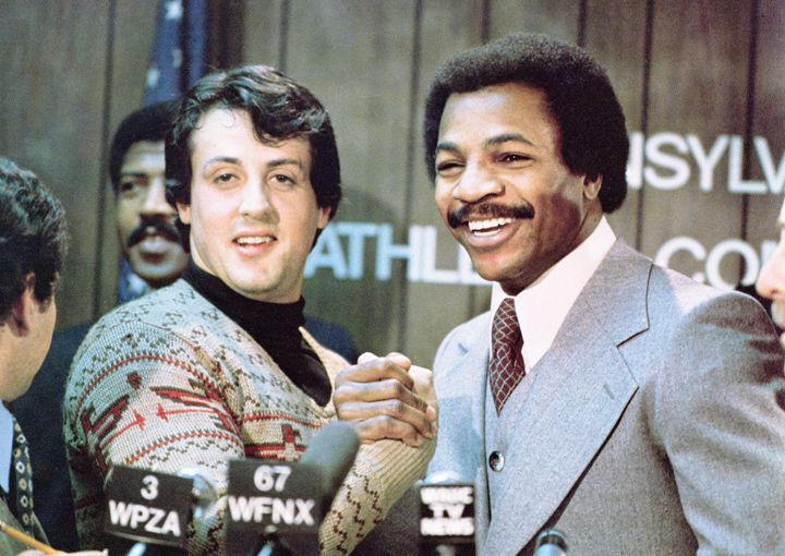 Sylvester Stallone and Carl Weathers grip hands and smile together during a press conference in a still from the film, "Rocky," directed by John G. Avildsen, 1976. (Photo by United Artists/Courtesy of Getty Images)