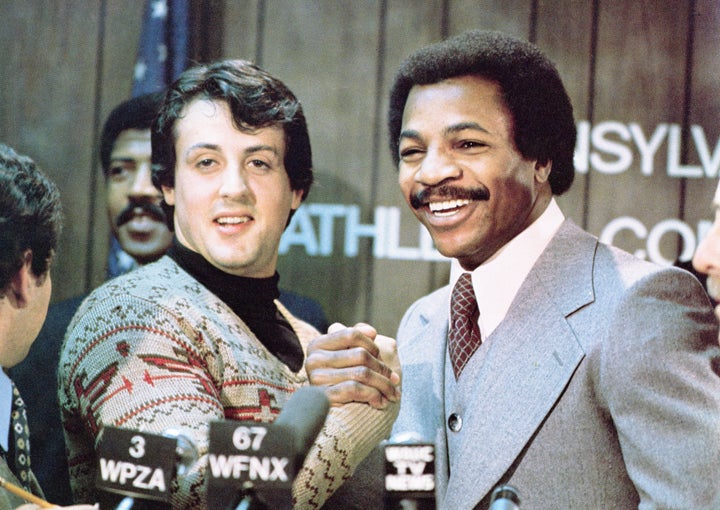 Sylvester Stallone and Carl Weathers grip hands and smile together during a press conference in a still from the film, "Rocky," directed by John G. Avildsen, 1976. (Photo by United Artists/Courtesy of Getty Images)