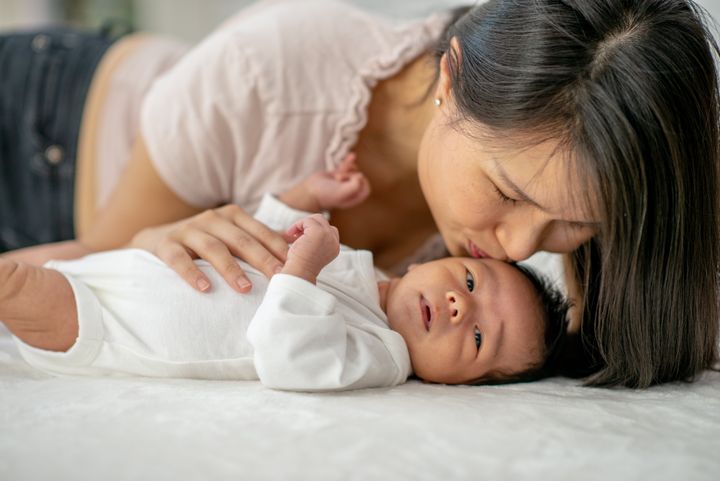 A new mother leans in to kiss her son's cheek. The newborn is laying on the bed in a onesie and is awake.