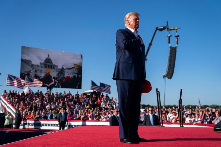 As footage from the Jan. 6, 2021, insurrection at the U.S. Capitol is displayed in the background, former President Donald Trump stands while a song, "Justice for All," is played during a campaign rally at Waco Regional Airport, on March 25, 2023, in Waco, Texas. The song features a choir of men imprisoned for their role on Jan. 6, 2021, singing the national anthem and a recording of Trump reciting the Pledge of Allegiance.