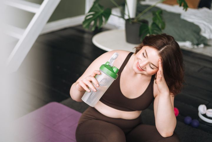 Young woman plus size in sports clothes training and drinking water in room at home