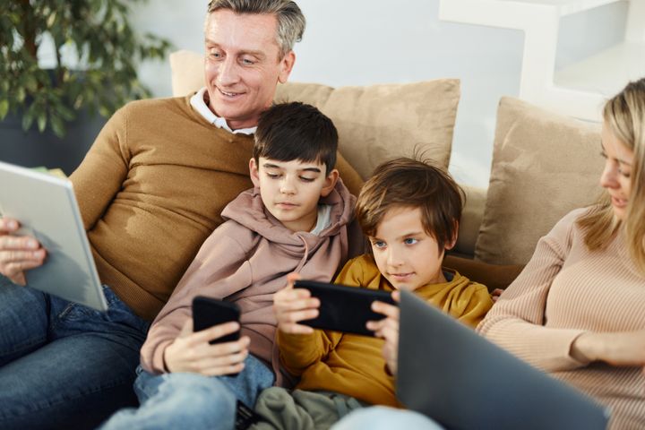 Happy parents and their small sons using wireless technology on sofa in the living room.