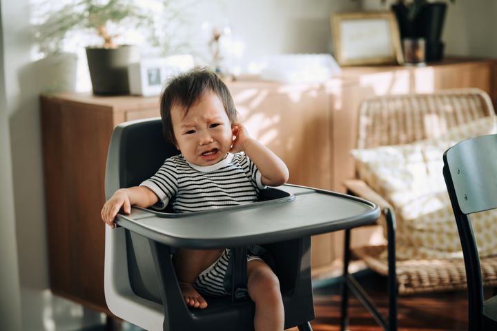 Portrait of crying Asian baby girl sitting on the high chair at home. Crying baby girl. Crying baby looking for attention and company