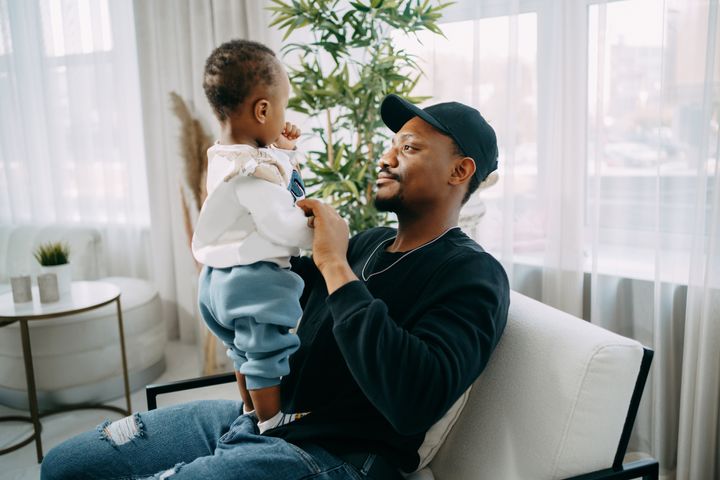 Black African dad in a cap in jeans relaxes while sitting in a chair with his little son after work in a cozy home