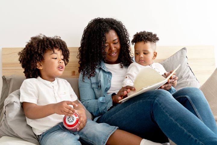Horizontal mid waist view of mother and children reading fairytales book relaxing sitting on bed at home. Family lifestyles