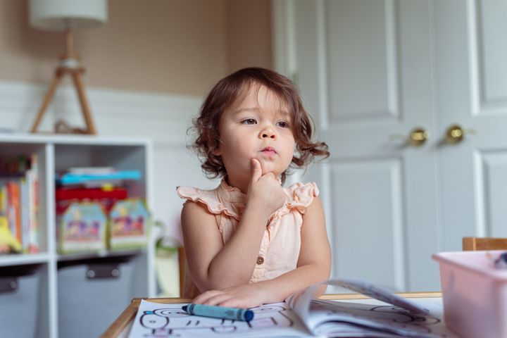 A cute preschool age girl looks off into the distance with a thoughtful and dreamlike expression while coloring and playing at home. Creativity, learning, curiosity and homeschooling concepts.