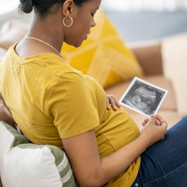 A pregnant woman sits on a sofa, holding out an ultrasound picture. While babies rightly receive attention and resources from our medical system, the people giving birth are sometimes abandoned.
