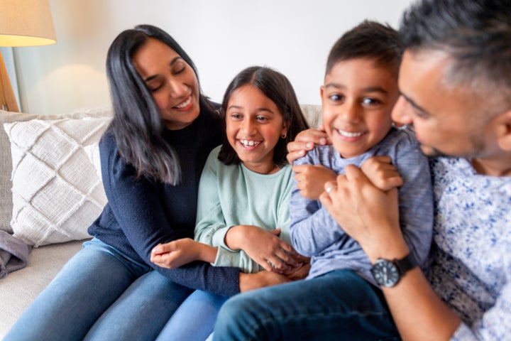 Loving having fun at home playing embracing and cuddling on the sofa. There is a boy and a girl child and 2 parents. They are happy, smiling and laughing