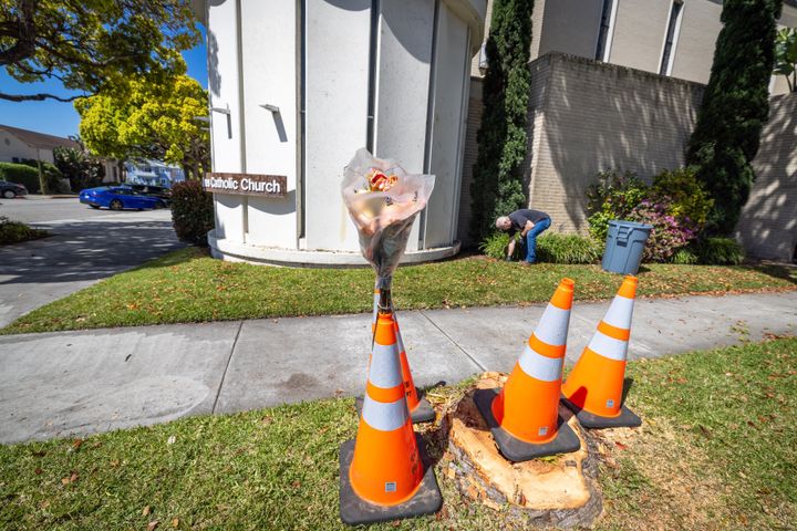 Redondo Beach, CA - April 09: Flowers are placed at the scene as Rob (no last name given), a custodial manager at Saint James Catholic Church, cleans up car parts on the grass where woman crashed her Porsche into a tree near the corner of PCH and Vincent Street in front of Saint James Catholic Church in Redondo beach, killing herself, this was after she killed her husband and left her two kids on the freeway. Police spent Monday following a bizarre and tragic trail of violence that left the dead and injured bodies of a Woodland Hills family scattered across Los Angeles County. The horror began with a woman killing her husband at their Valley home, abandoning her two small daughters on the 405 Freeway one of whom died and then fatally crashing her car into a tree in Redondo Beach, according to a law enforcement source who was not authorized to speak publicly about the investigation. Photo taken in in Redondo Beach Tuesday, April 9, 2024. (Allen J. Schaben / Los Angeles Times via Getty Images)