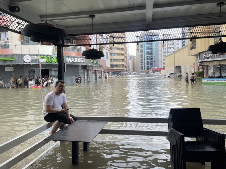 DUBAI, UNITED ARAB EMIRATES - APRIL 17: A man sits on the fence in front of a flooded store as people wade through submerged streets at flooded area while heavy rains negatively affect daily life in Dubai, United Arab Emirates on April 17, 2024. Due to heavy rains water puddles formed on the roads and vehicles were stranded on the roads in Dubai city. (Photo by Stringer/Anadolu via Getty Images)