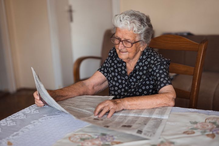 Serene senior lady with her eyeglasses reading newspapers while resting at her cozy home table